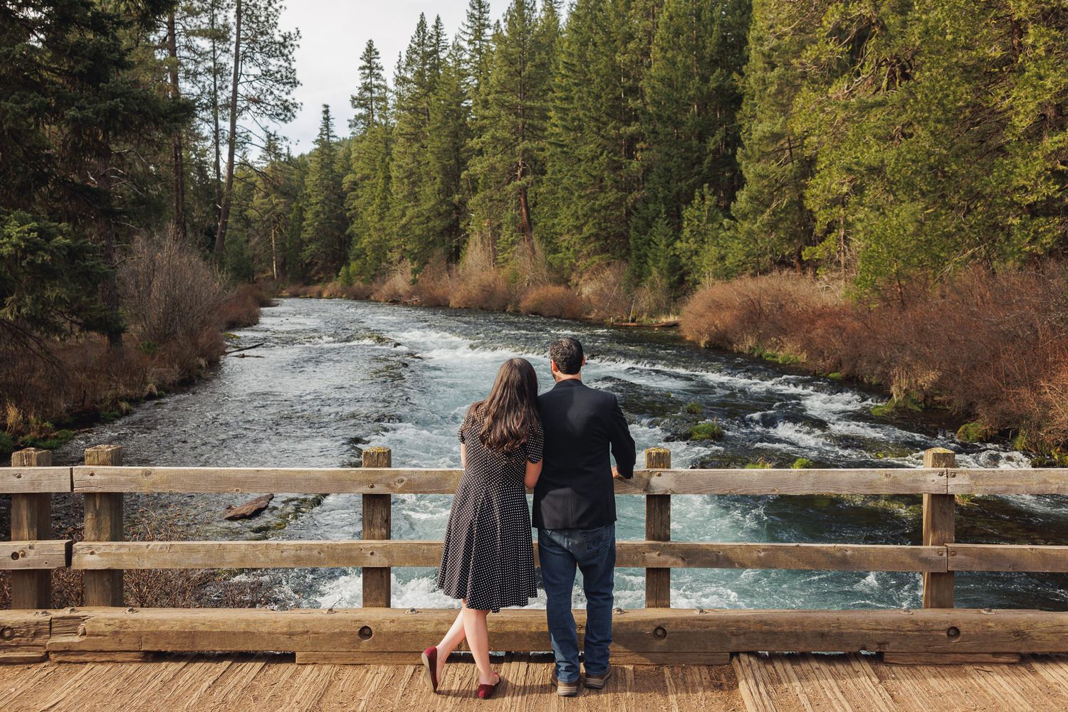 A couple standing on a wooden bridge overlooking a scenic river surrounded by pine trees.