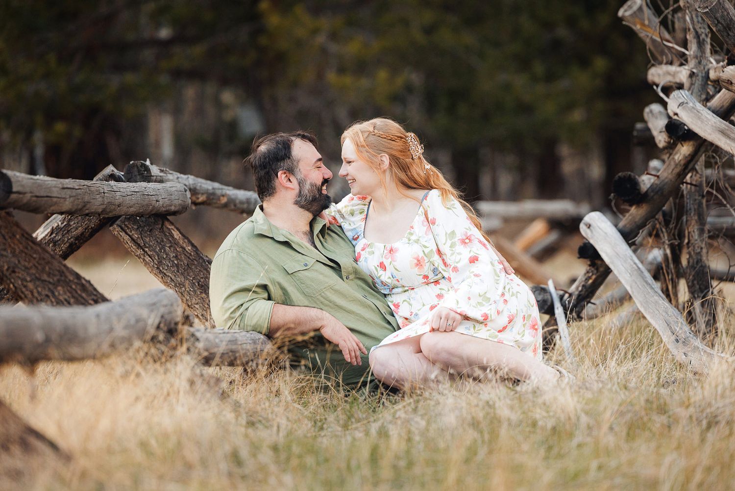 A couple sits in golden grass near rustic wooden fences, sharing a romantic moment outdoors.