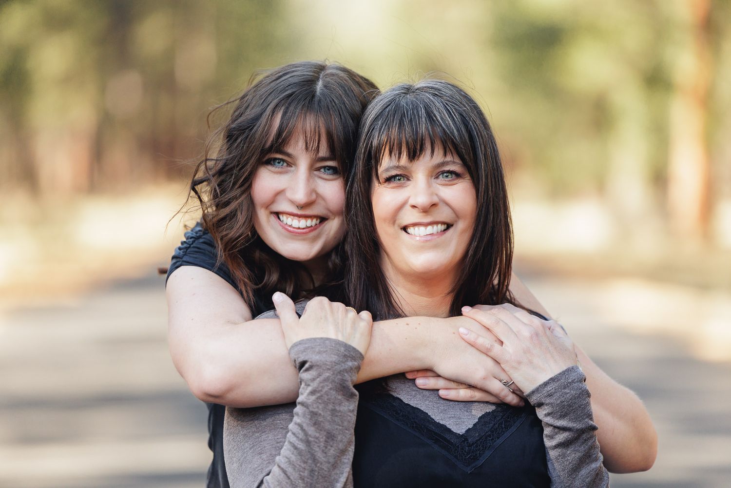Two smiling women with dark hair and bangs embrace warmly outdoors, one hugging the other from behind on a tree-lined path.