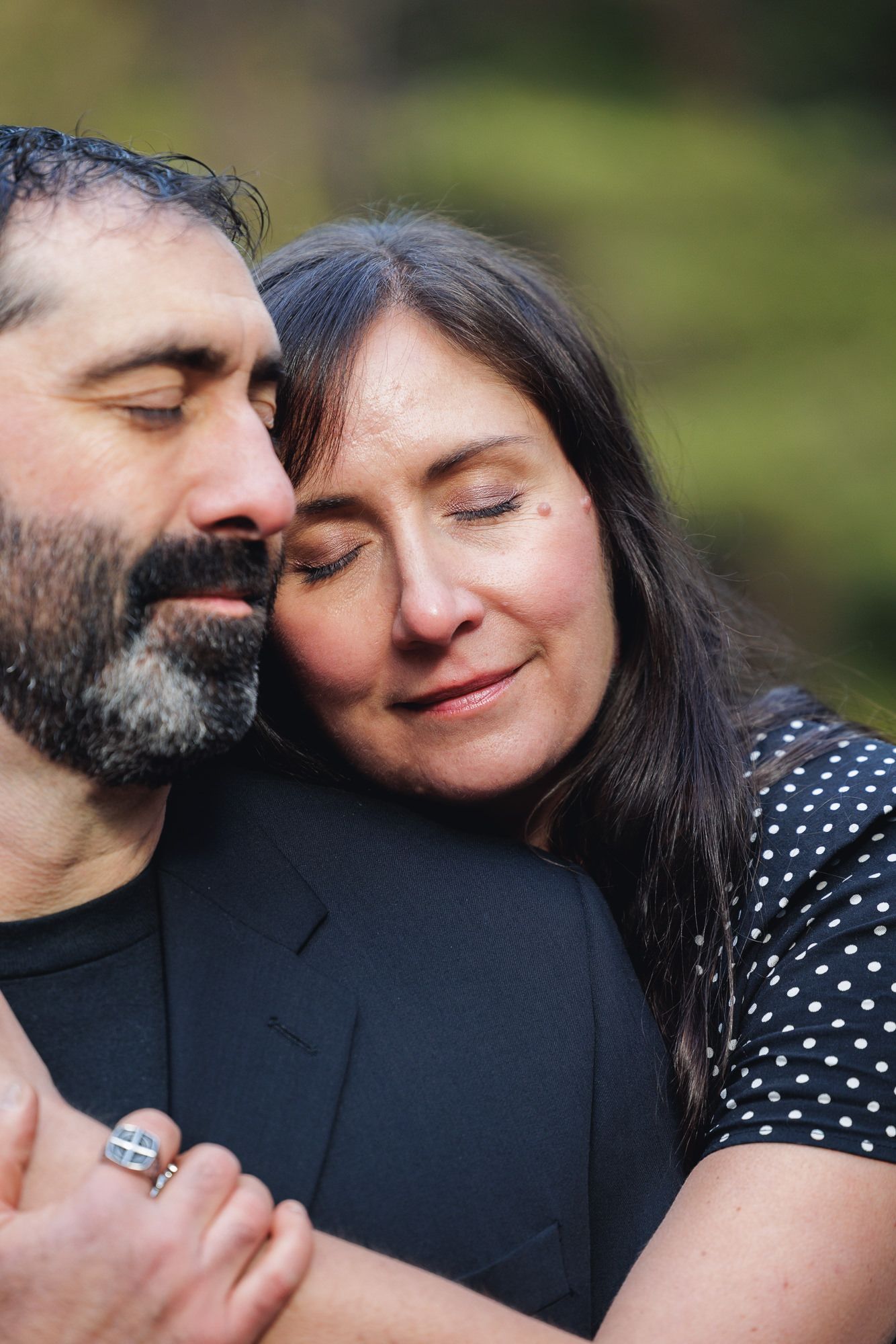 A couple with eyes closed share a tender embrace outdoors, woman resting her head on mans shoulder.
