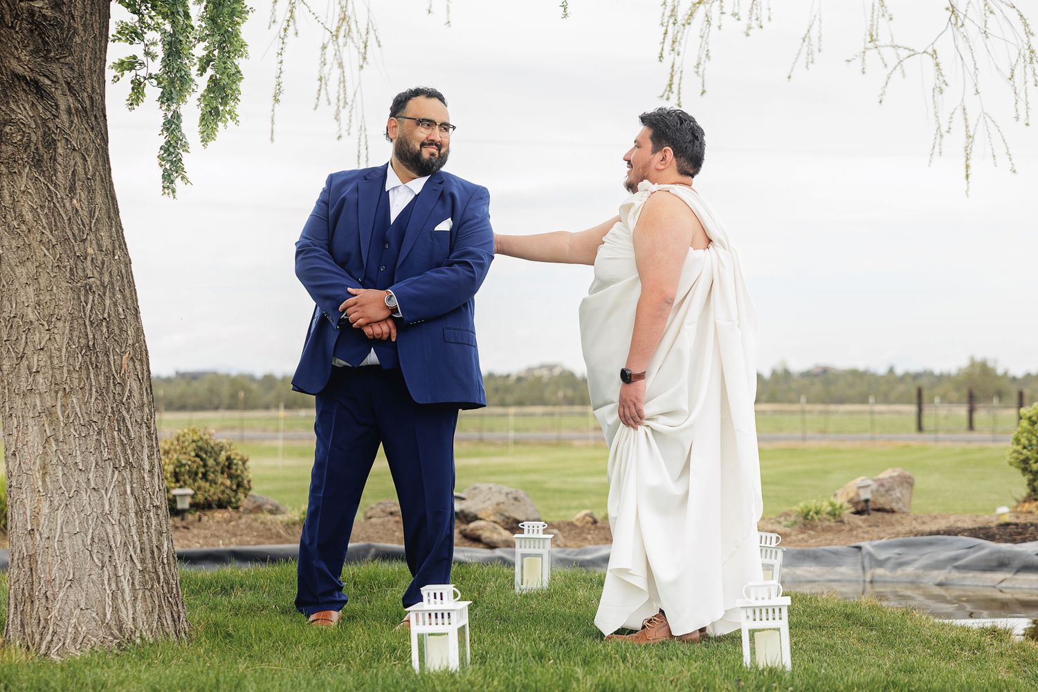 Two grooms share an emotional first look moment outdoors at a wedding venue, one in a blue suit, one in white.