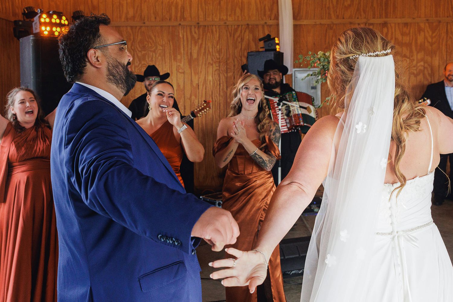 Bride and groom share first dance at rustic wedding reception with bridesmaids in rust dresses cheering behind them.