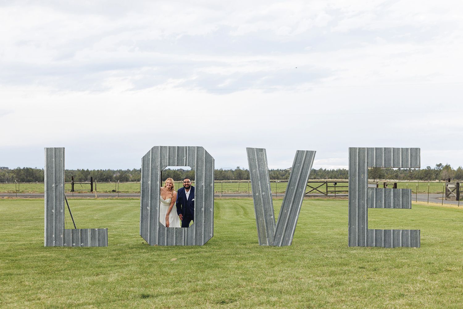 Couple poses inside giant metal LOVE letters on green lawn at outdoor wedding venue under cloudy sky.