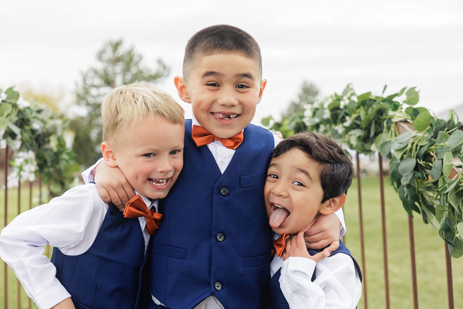 Three young boys in blue vests and orange bow ties smiling and hugging outdoors at a wedding.