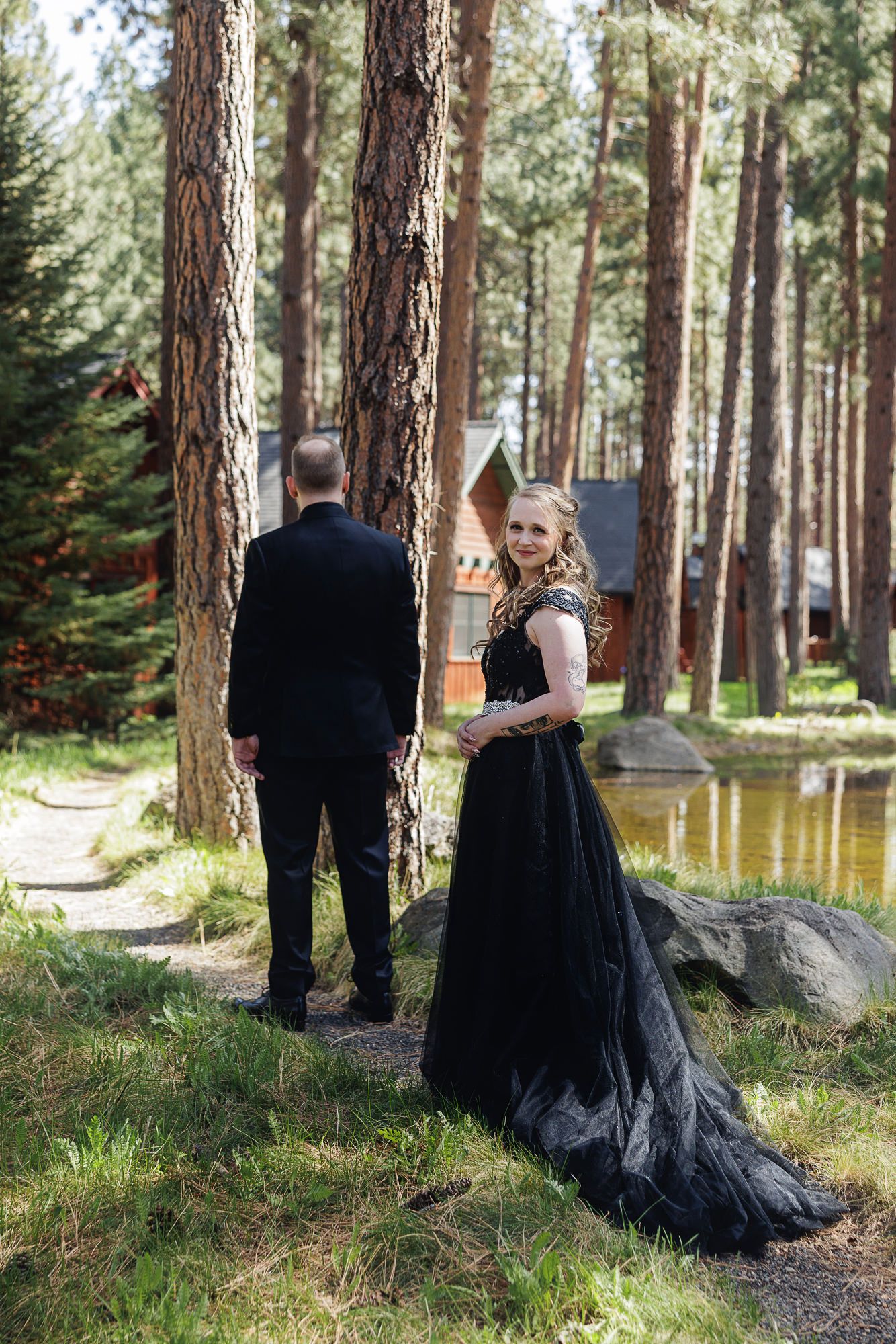 Couple in black formal wear posing in pine forest near pond, woman in elegant black gown with train looks back at camera.