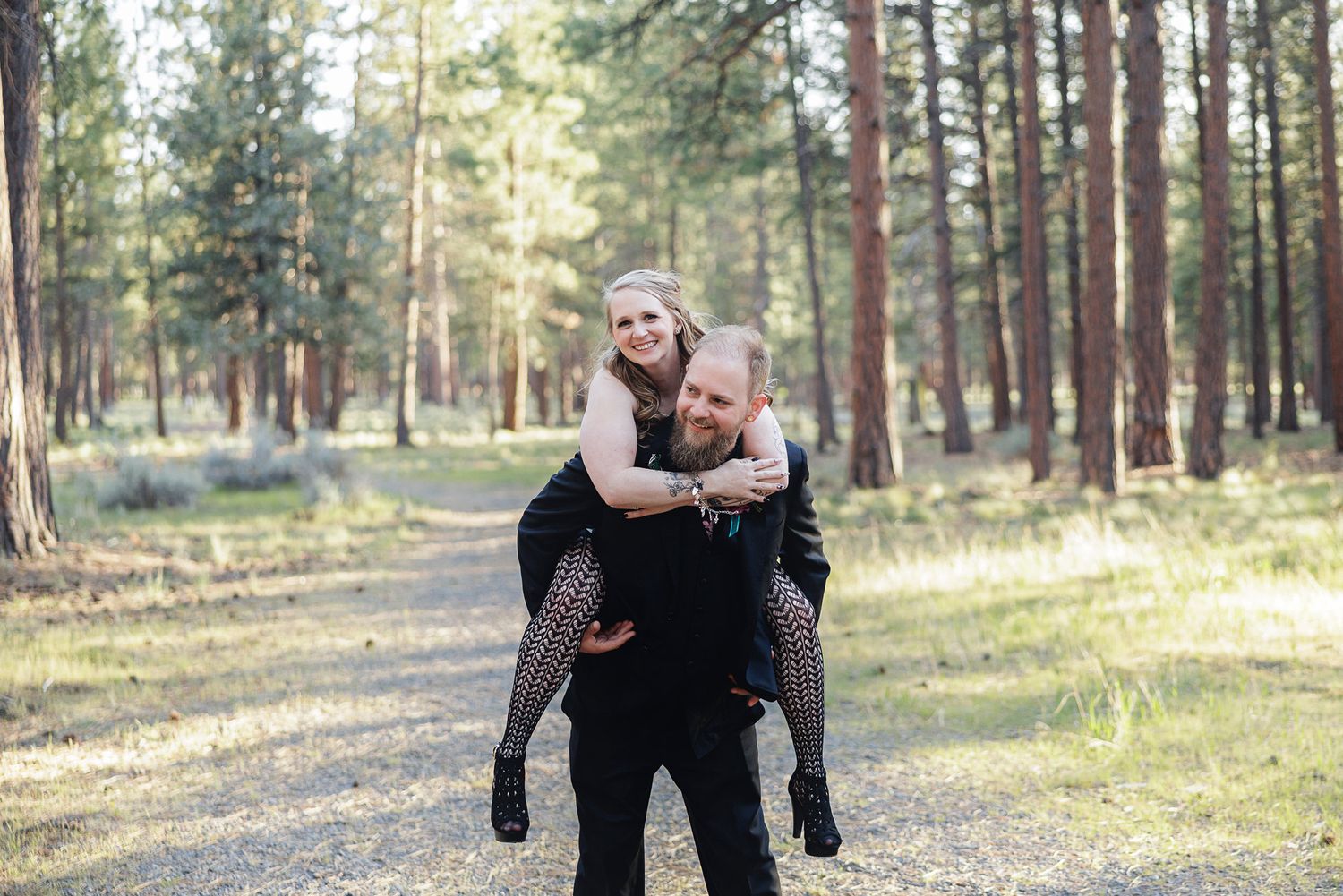 A man giving a woman a piggyback ride on a forest trail, both smiling, surrounded by tall pine trees.