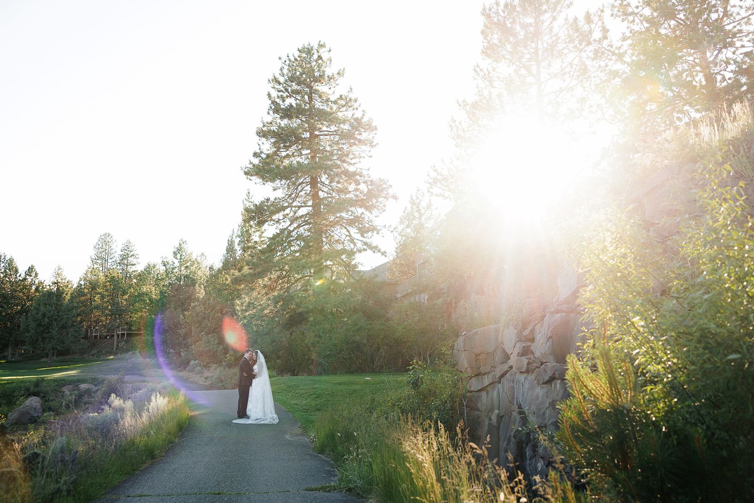 Bride and groom share a romantic moment on a sunlit path surrounded by lush trees and greenery at an outdoor wedding.
