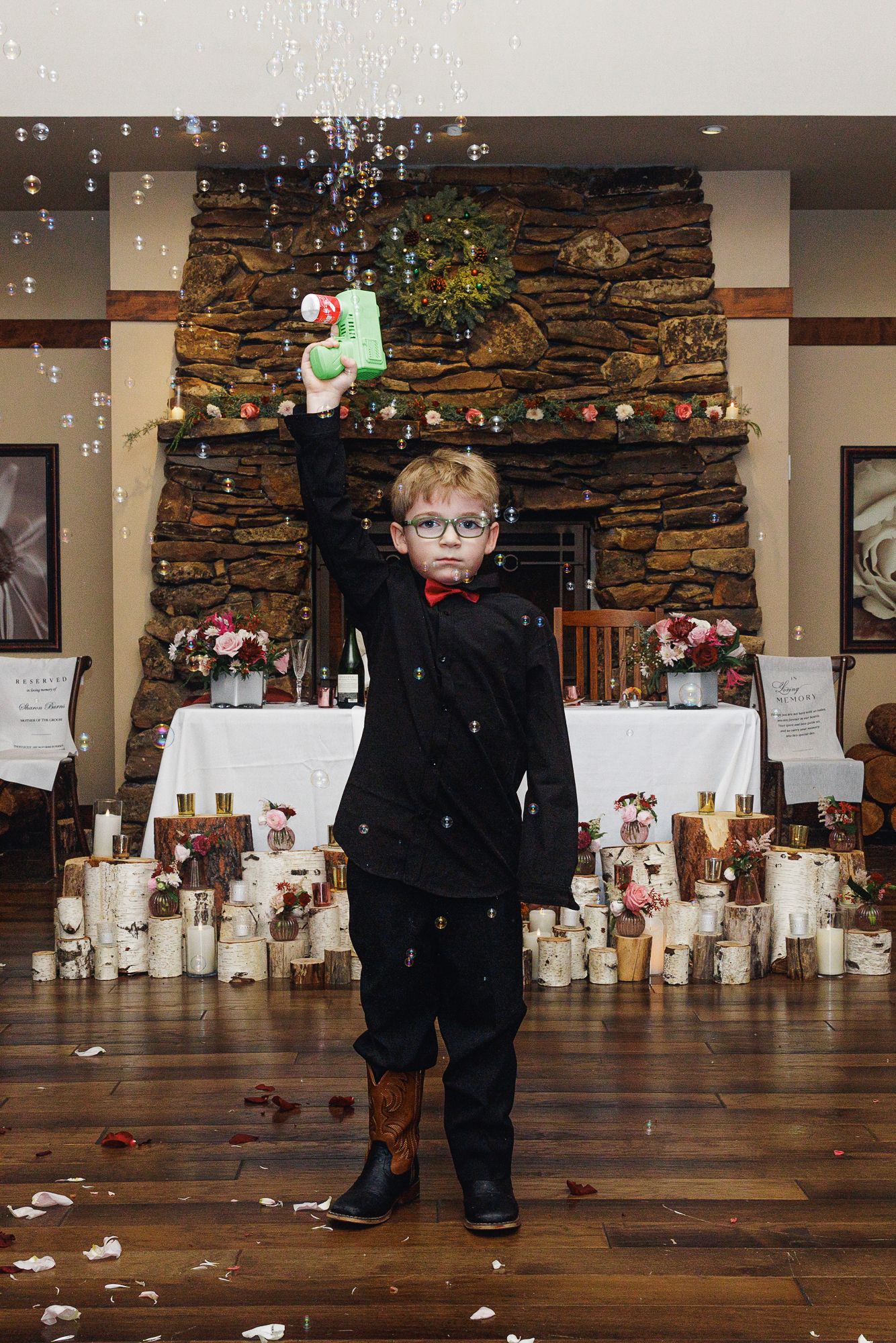 A young child in formal black attire stands alone in front of a stone wall backdrop with white candles on the floor.