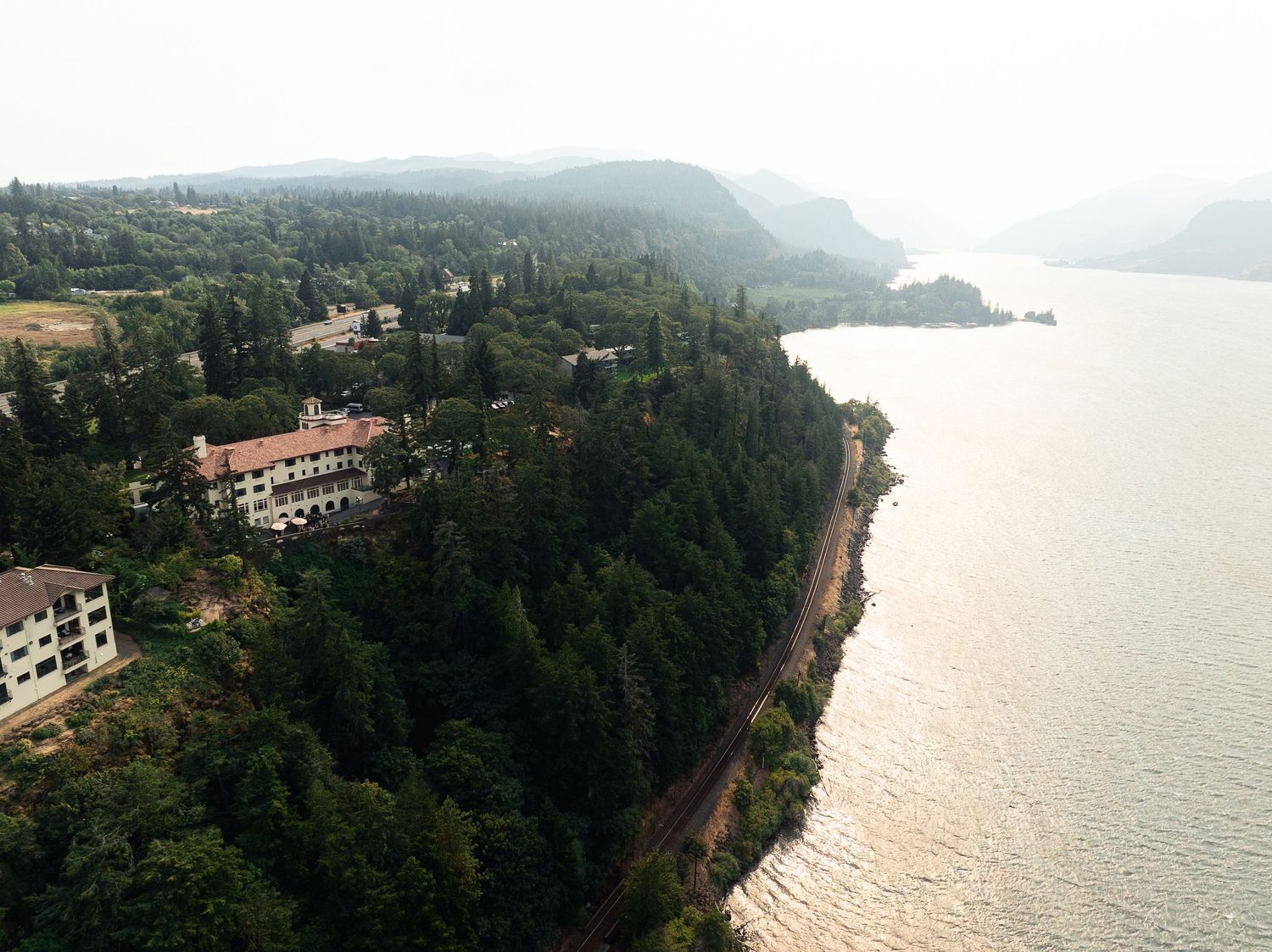 Aerial view of a forested Columbia River Gorge shoreline with historic lodge buildings nestled among tall evergreen trees.