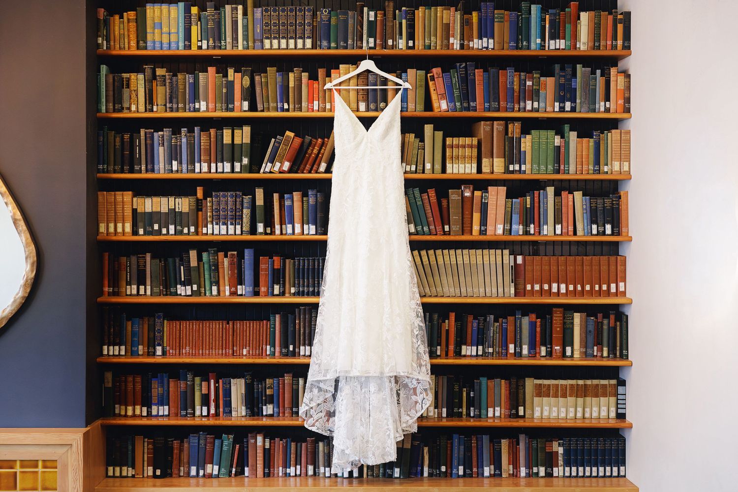 Elegant lace wedding dress hanging on a hanger in front of a wooden bookshelf filled with colorful books.