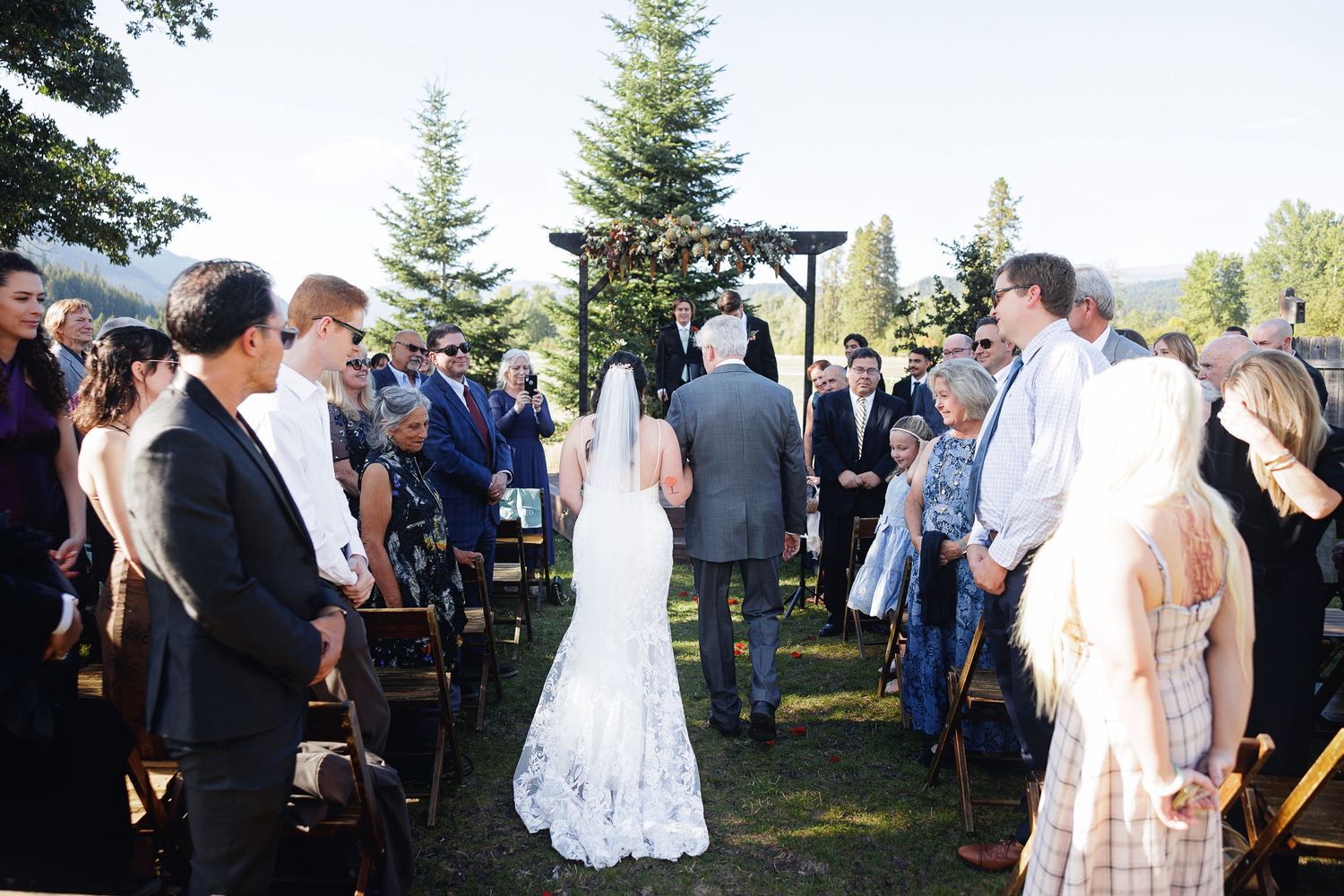 Bride walking down the aisle at an outdoor wedding ceremony surrounded by guests on a sunny day.