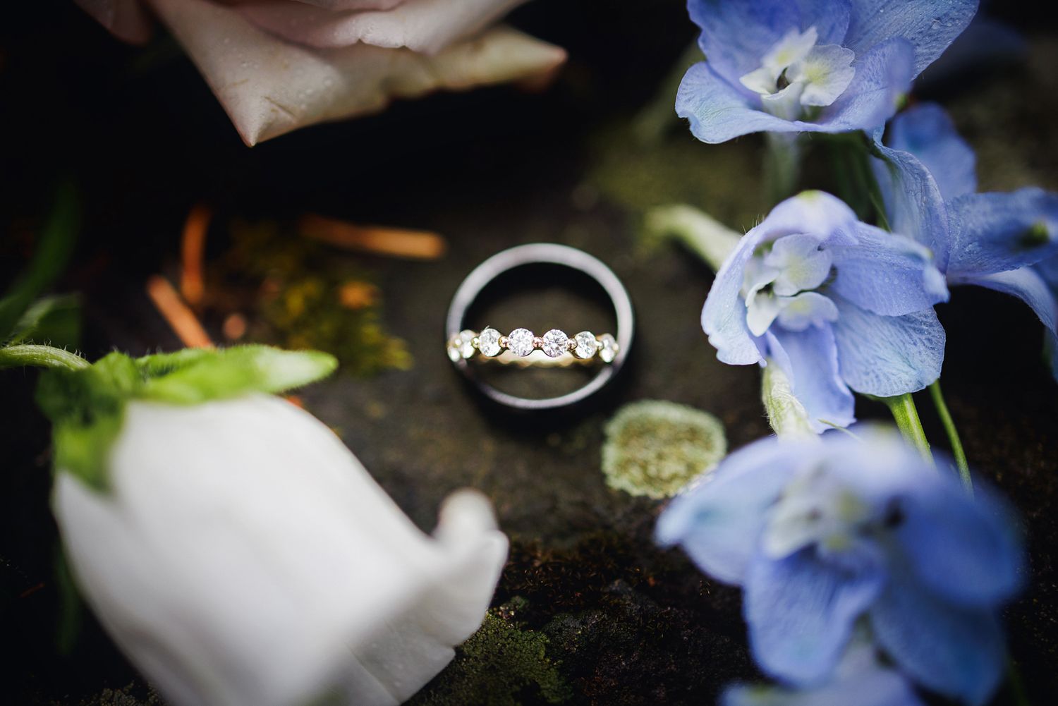 Wedding rings photographed with delicate blue hydrangea flowers and white decor against a dark background.