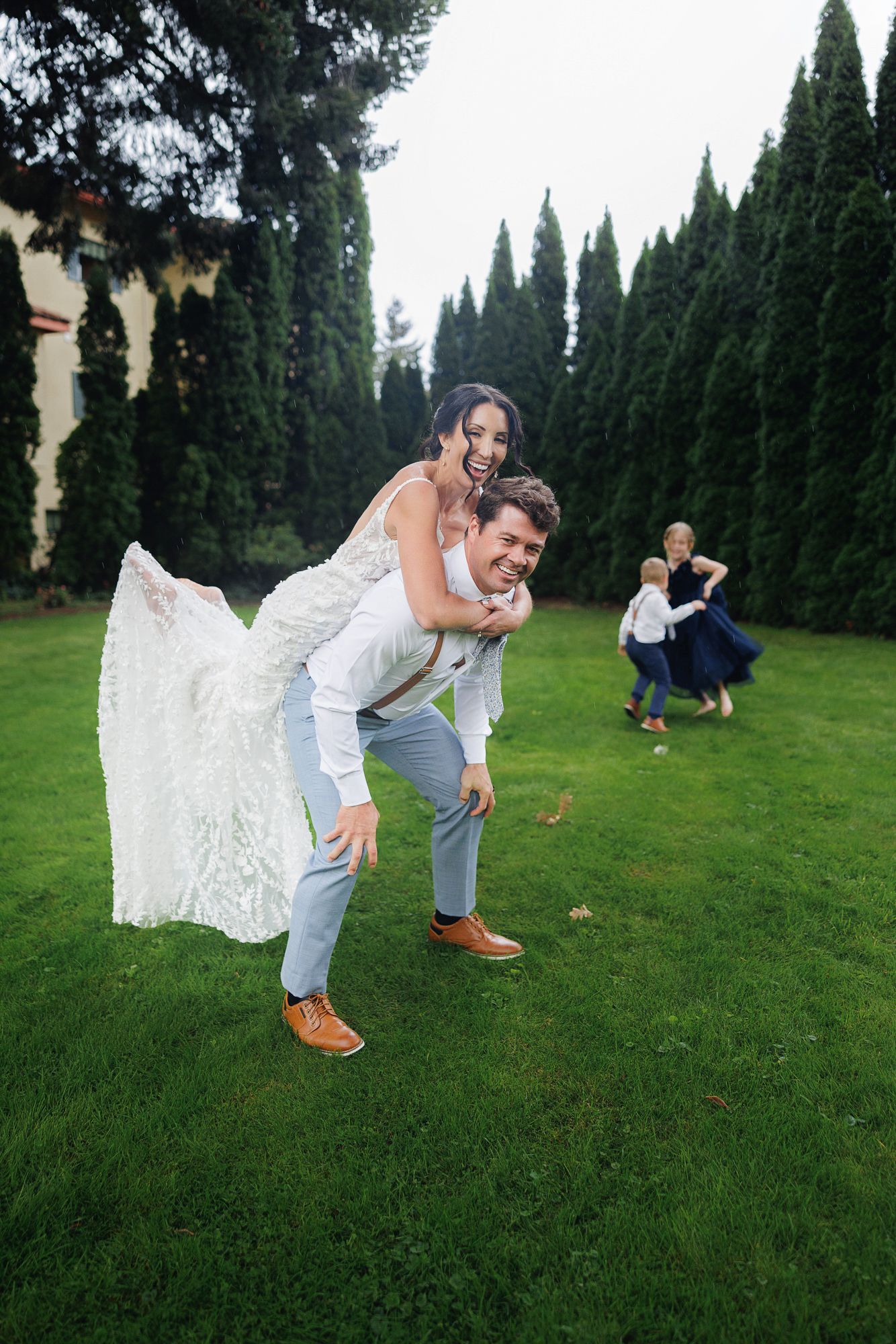A wedding couple performs a coordinated dance move on grass with tall cypress trees creating a dramatic backdrop.