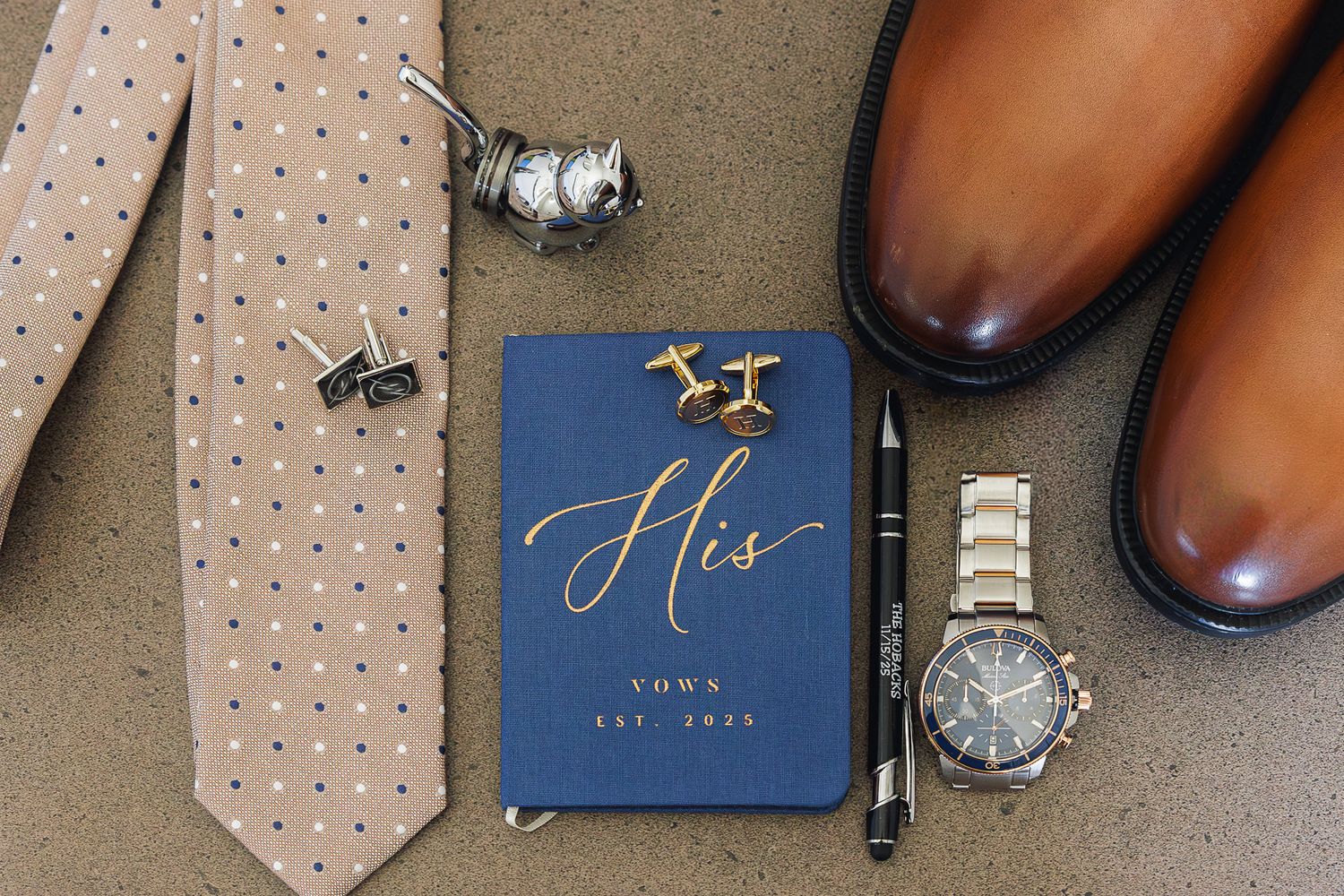 Wedding accessories including a polka dot tie, blue vow book, brown leather shoes, cufflinks and wristwatch on grey surface.
