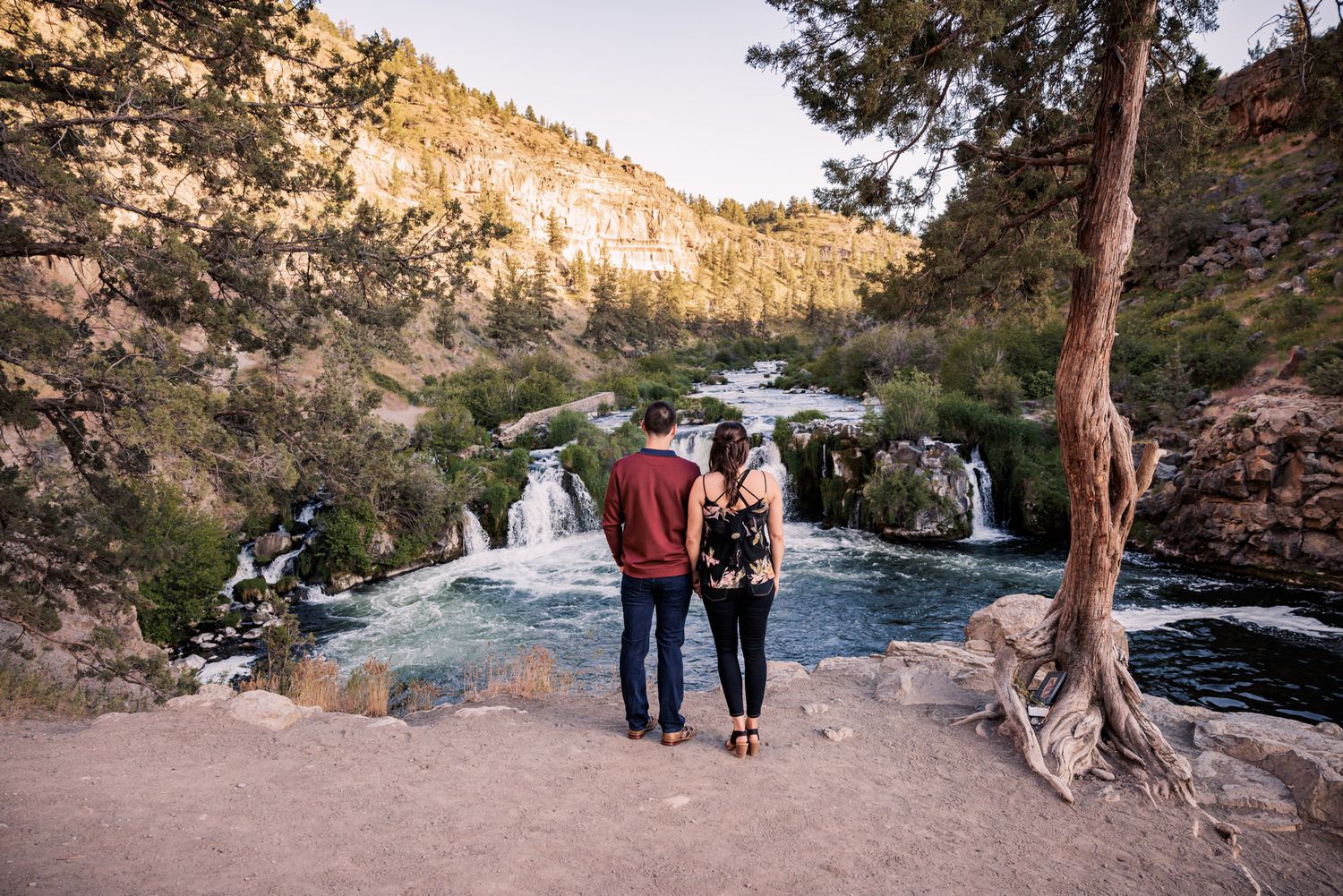 Two people walk hand in hand along a scenic mountain trail with waterfalls and rocky cliffs in the background.