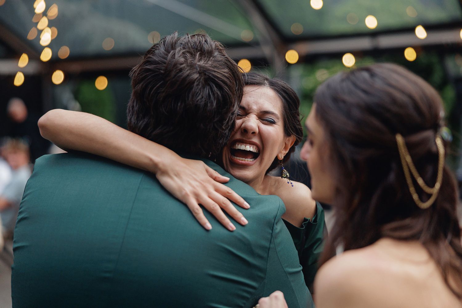 Woman laughing joyfully while hugging a man in green at an outdoor evening celebration with string lights.