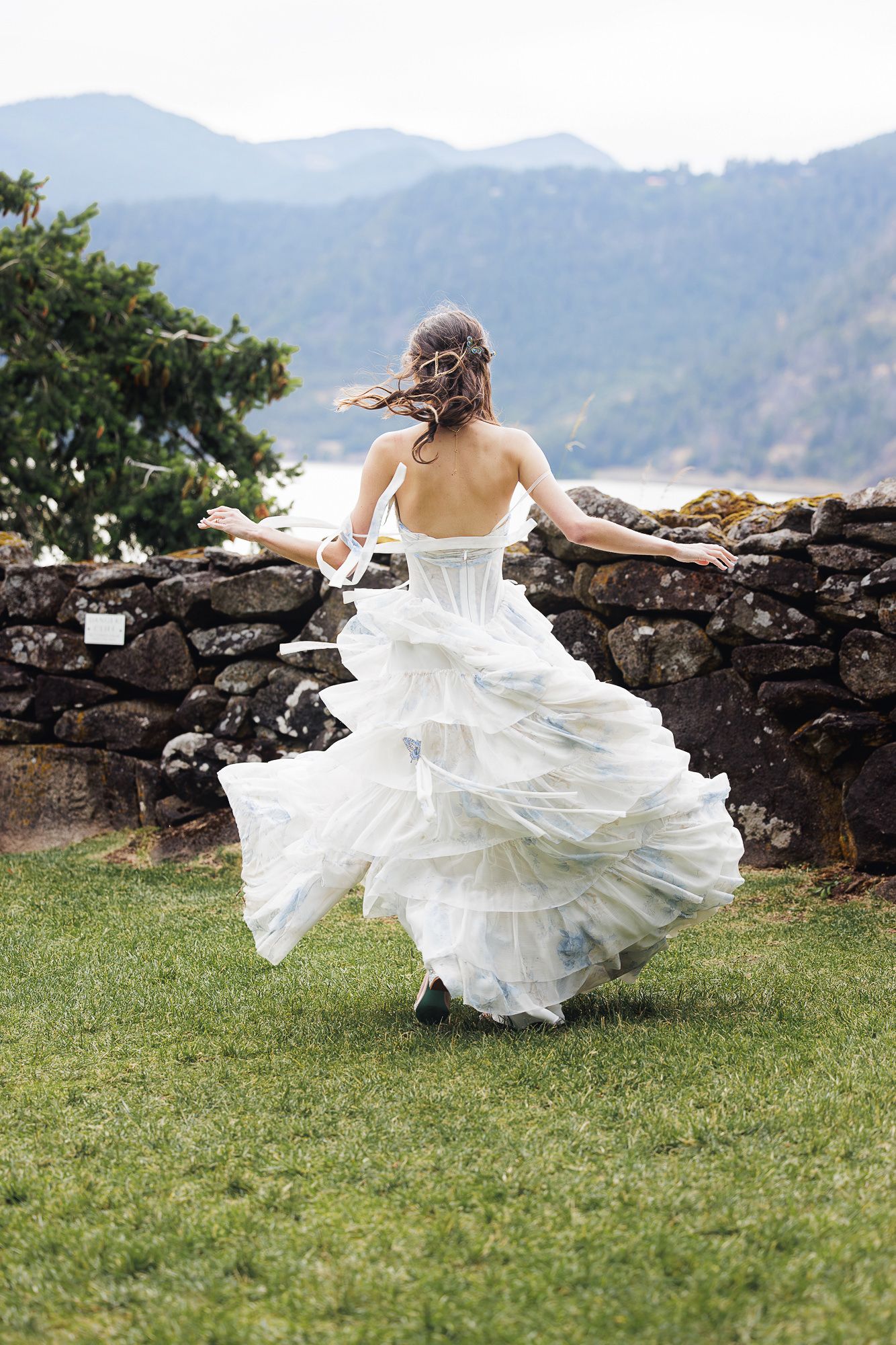 Bride in flowing white wedding gown running across green grass toward stone wall with mountain lake backdrop.
