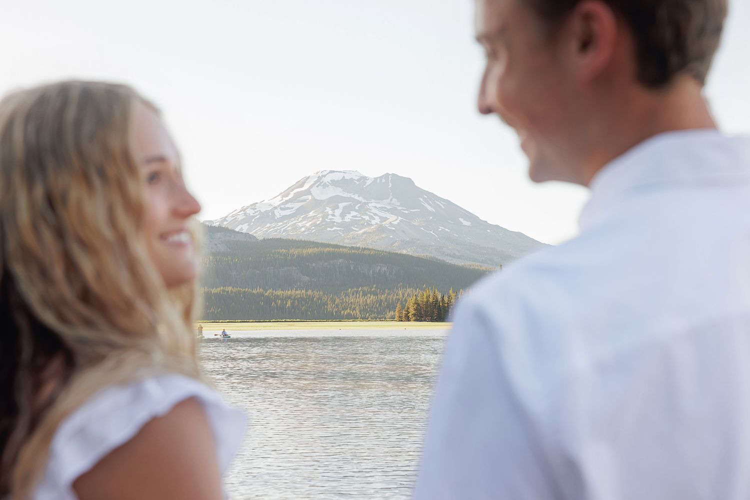 Couple smiling at each other with a scenic mountain lake and snow-capped peak in the background.