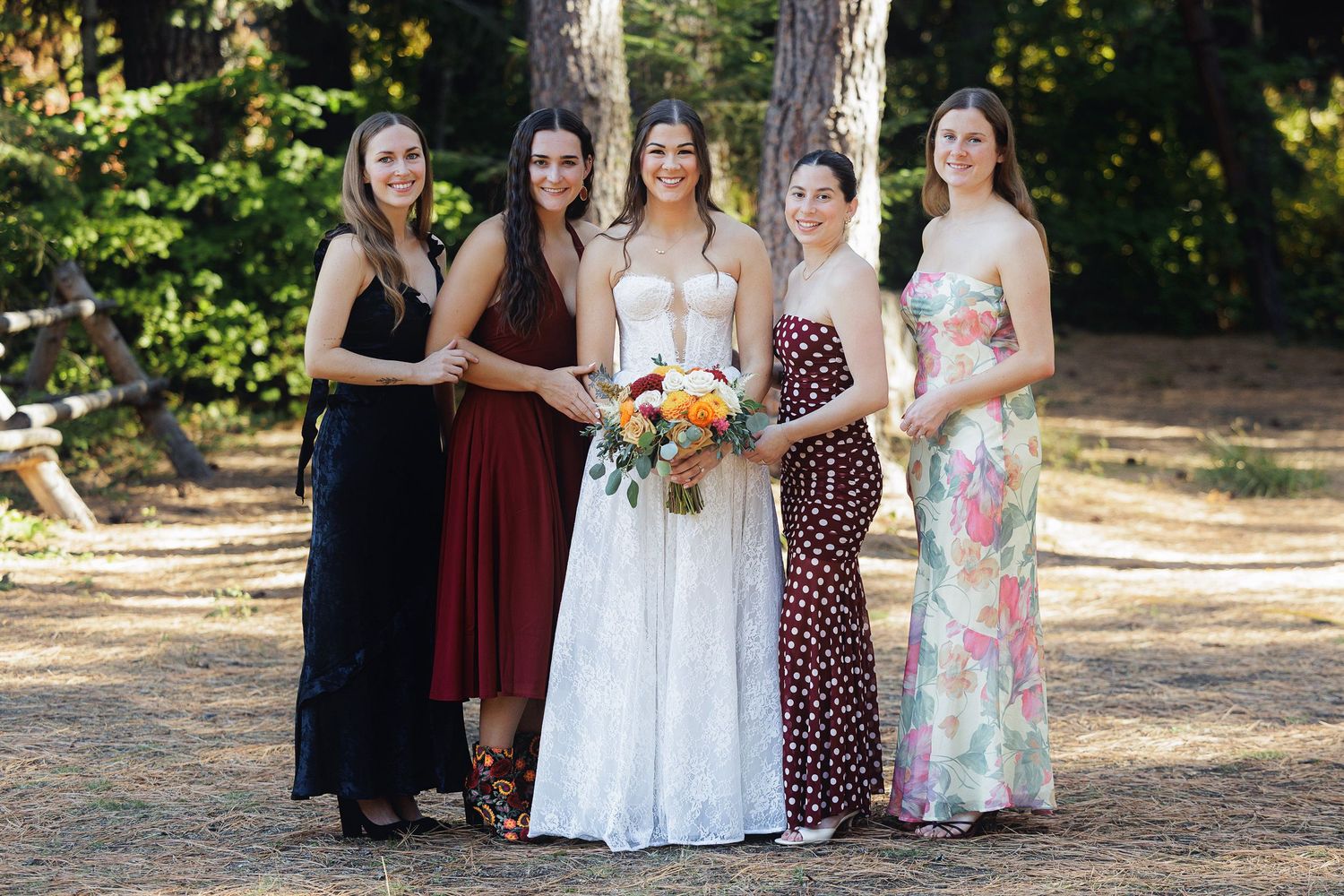 Bride in white gown poses with four bridesmaids in colorful dresses outdoors among trees.