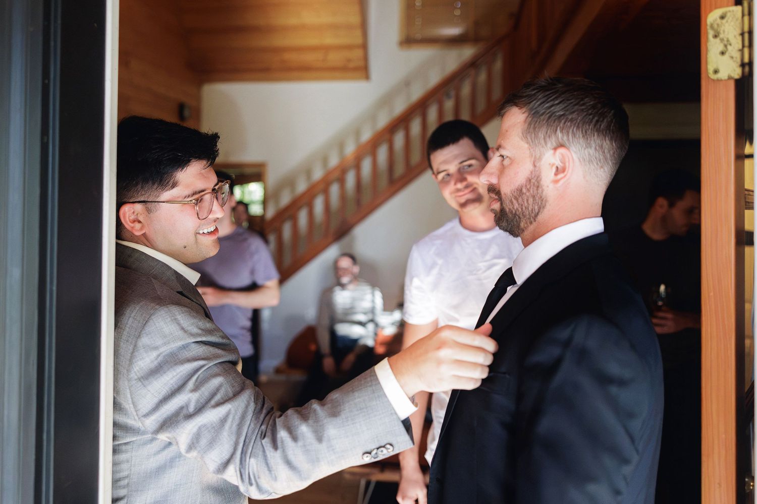 Groomsmen helping groom adjust tie before wedding ceremony indoors near staircase.