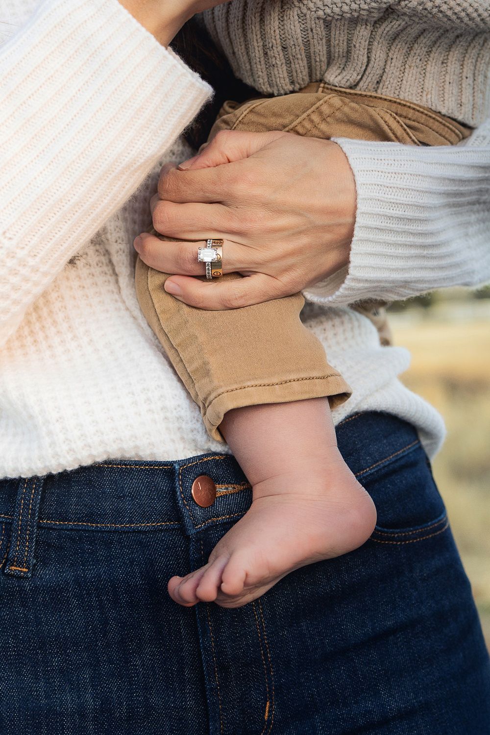 A baby's tiny bare foot rests against denim jeans while hands wearing a diamond ring embrace the little one.