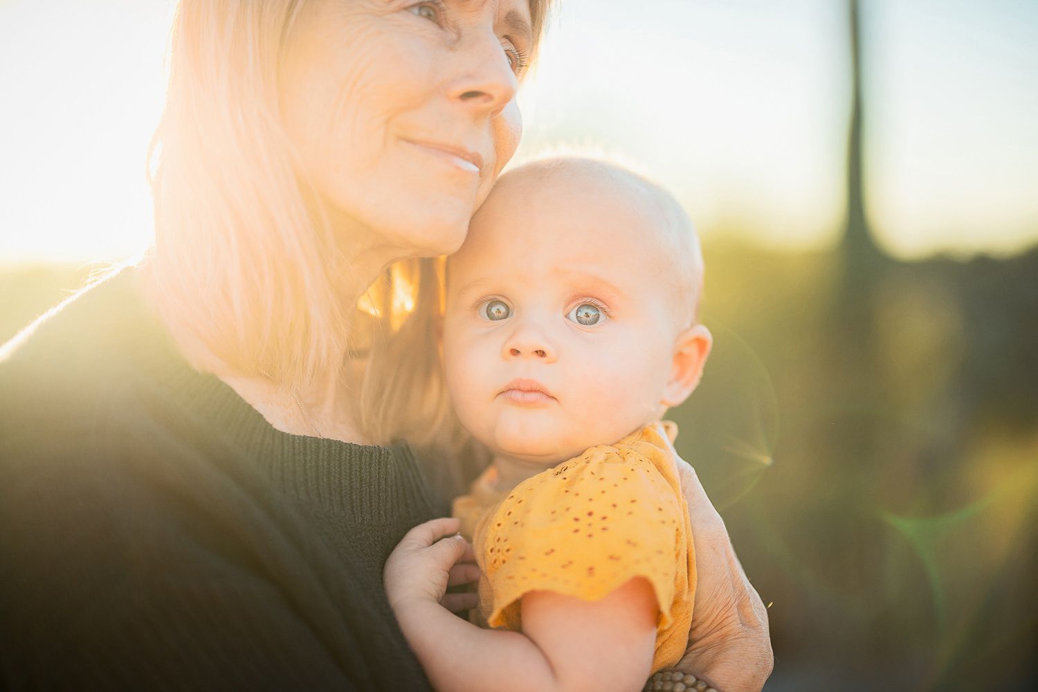 A baby in a yellow onesie is held in sunlit embrace during golden hour.