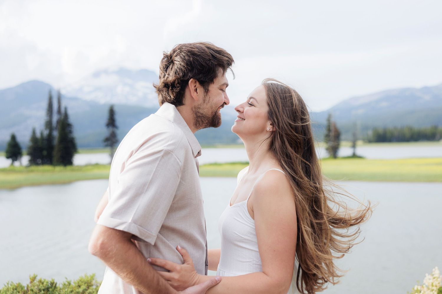 A romantic couple embraces against a scenic mountain backdrop with a lake and evergreen trees in the distance.