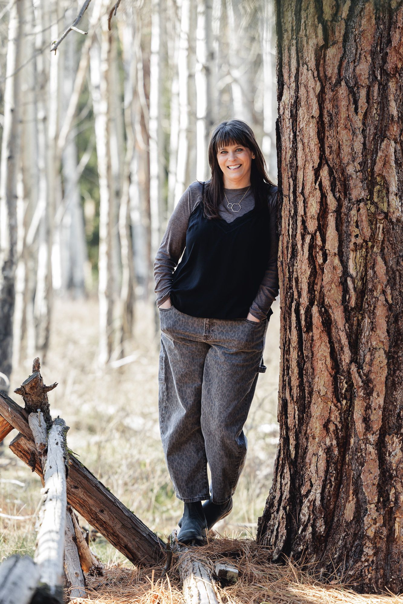 Smiling woman in gray outfit leaning against a large pine tree in a birch forest.