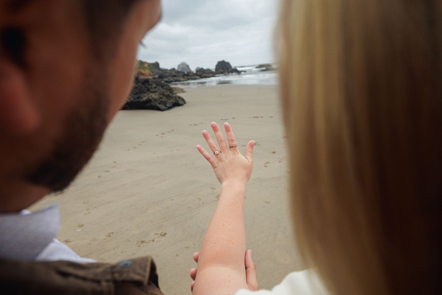 Couple on beach admiring engagement ring, ocean and rocky shore in background.