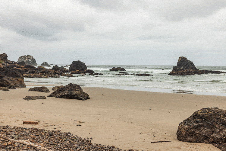 Rocky beach with large boulders on sandy shore, ocean waves and sea stacks under overcast sky.