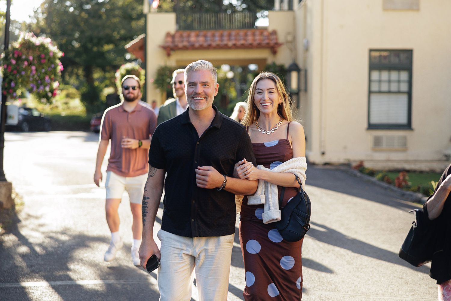 A smiling couple walks arm-in-arm outdoors near a building, with two people following behind on a sunny day.