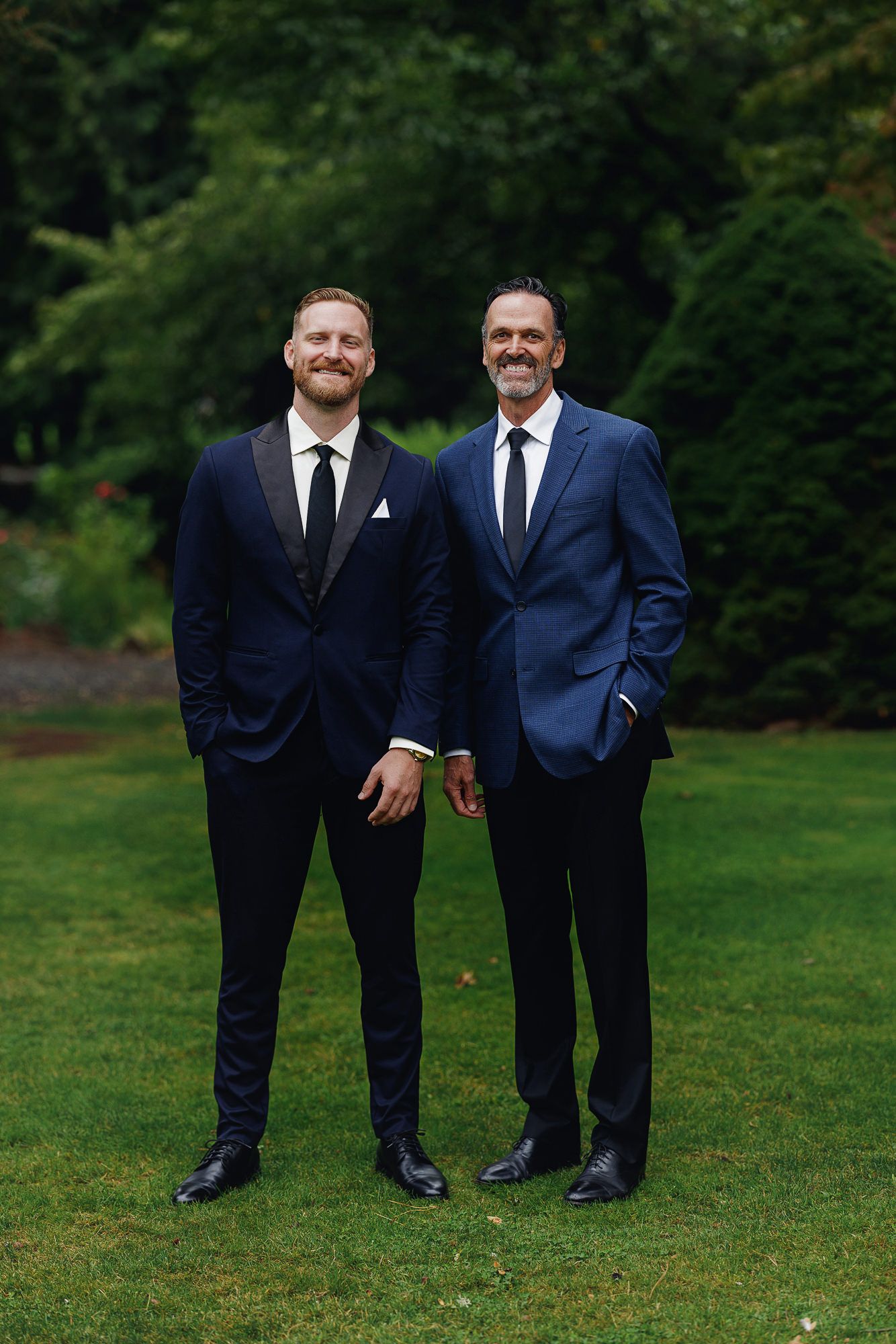 Two men in navy suits smile while standing on green grass with lush trees in the background at a wedding.