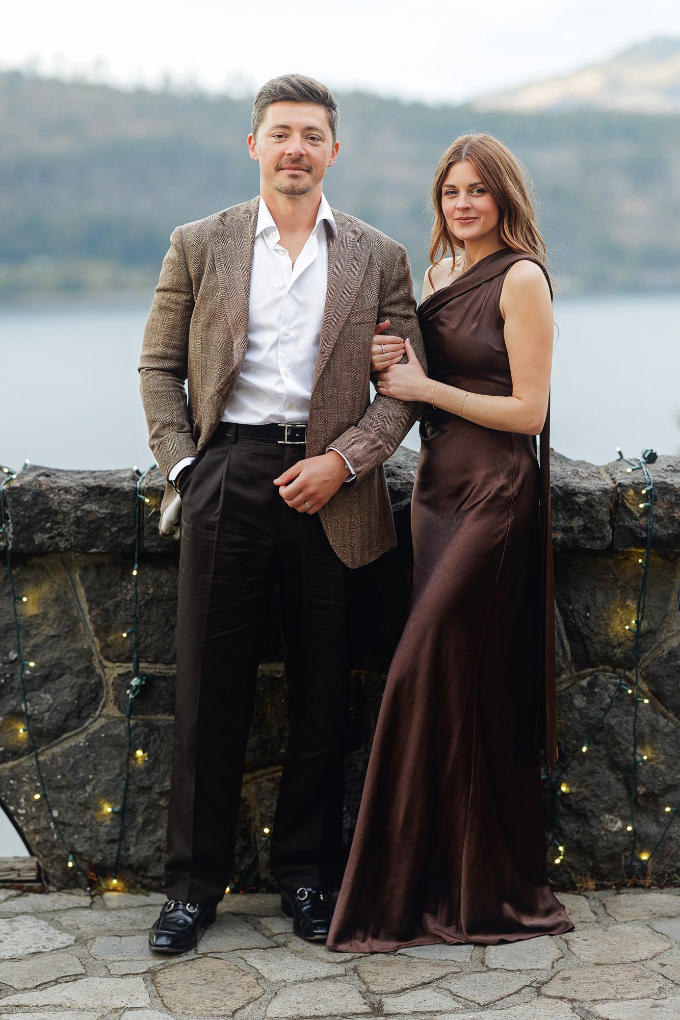 Elegant couple posing outdoors, man in brown blazer and woman in satin gown, with scenic lake and mountains behind.