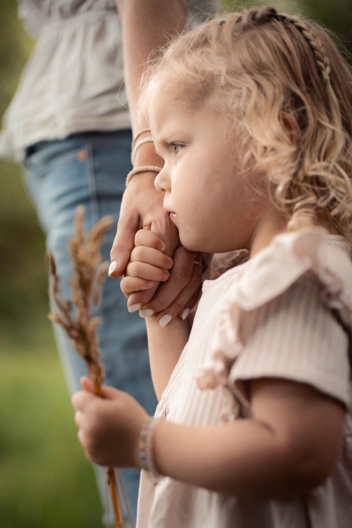 A young child in a light dress holds a branch while kissing someone's hand during a tender outdoor moment.