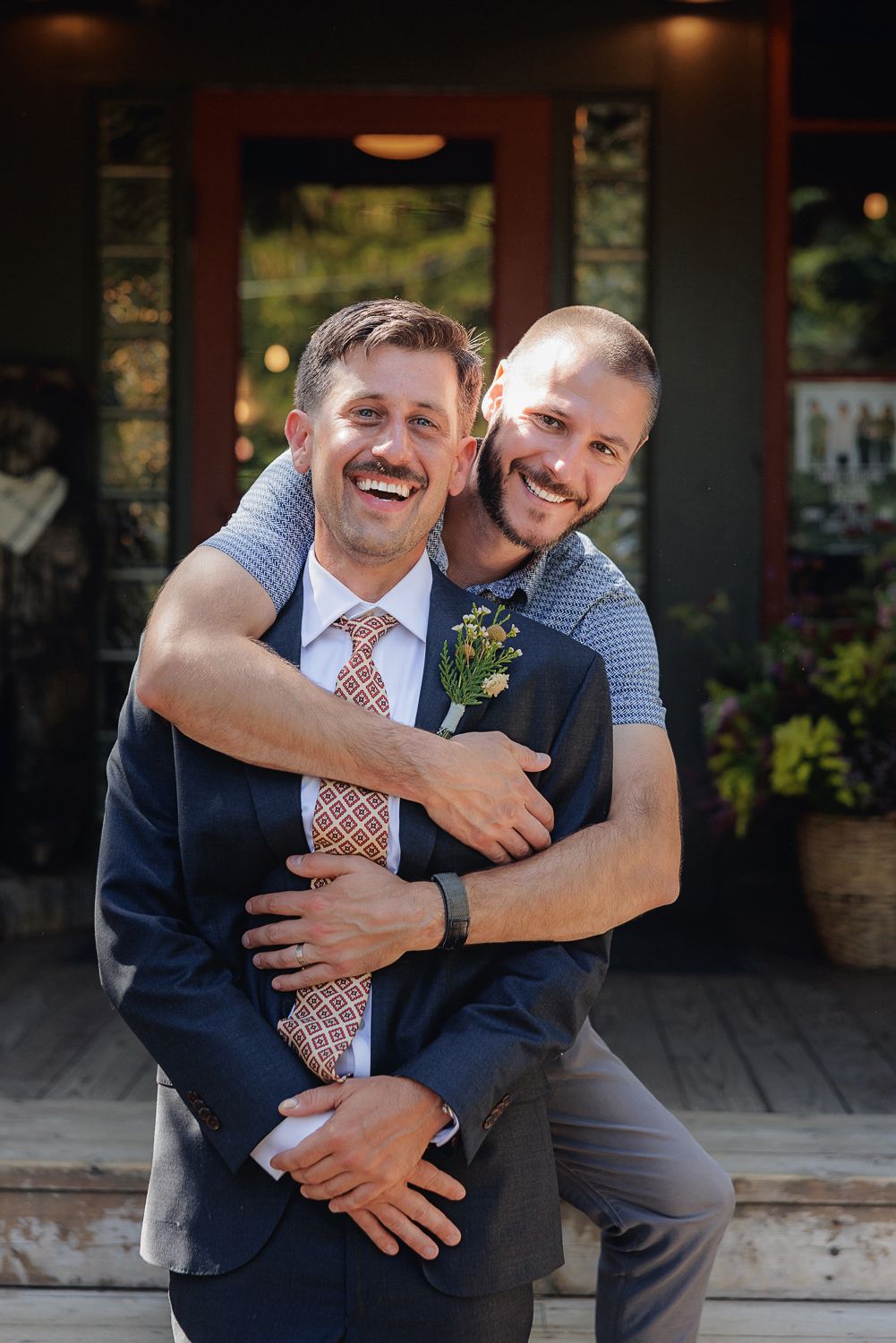 Two people embrace and laugh together on a porch, one in a navy suit and one in casual blue attire.