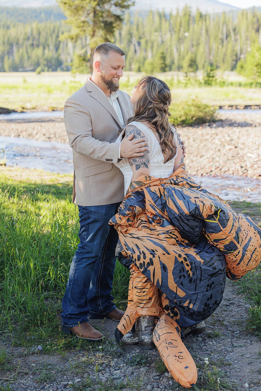 A couple shares a romantic dance pose along a riverside with mountains and forest in the background.
