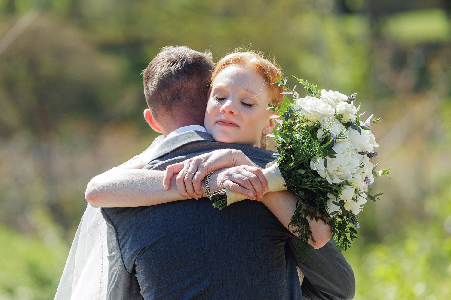 Bride with red hair hugging groom in suit, holding white floral bouquet outdoors on sunny wedding day.