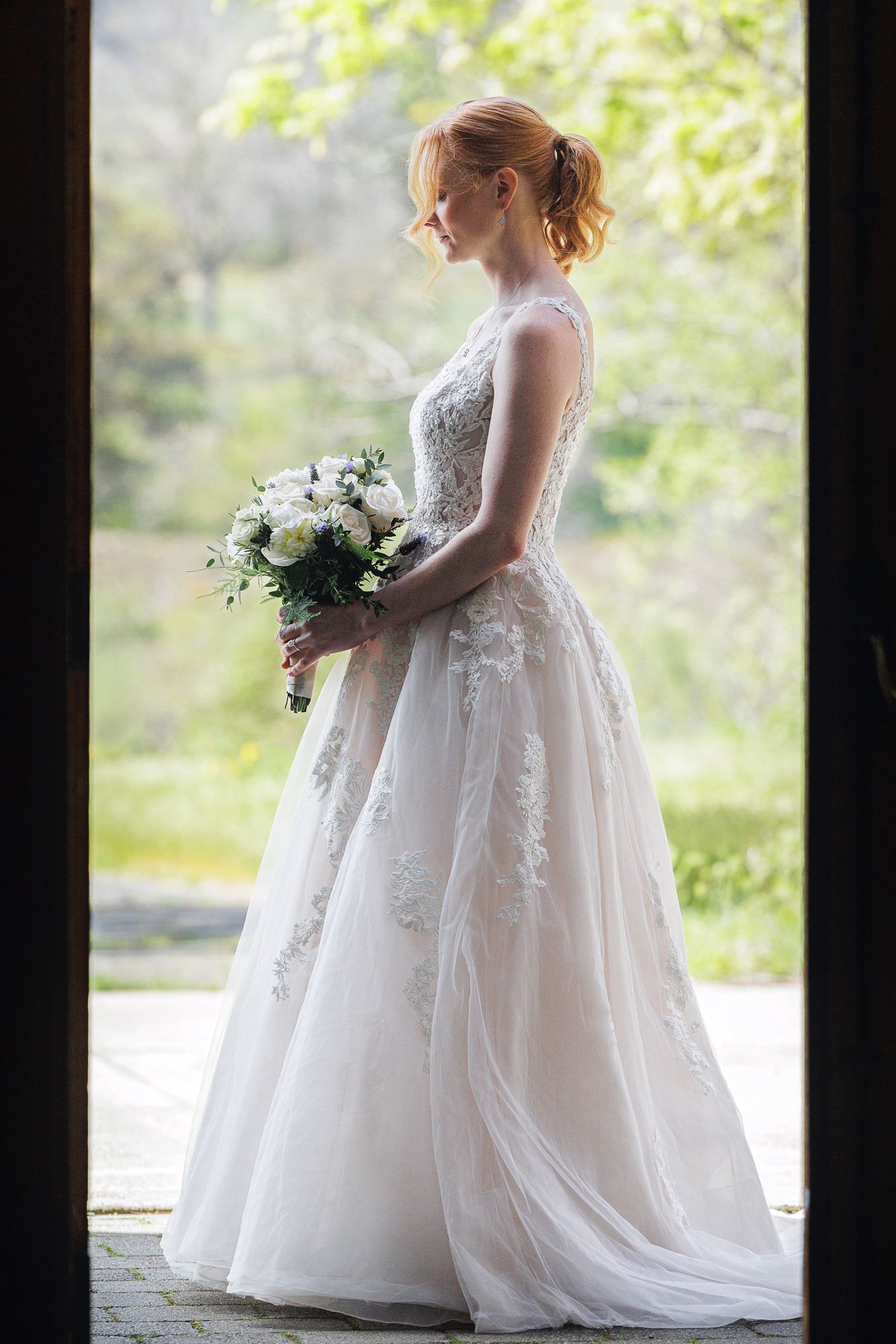 Elegant bride in lace wedding gown holding white floral bouquet, standing in doorway with green nature backdrop.