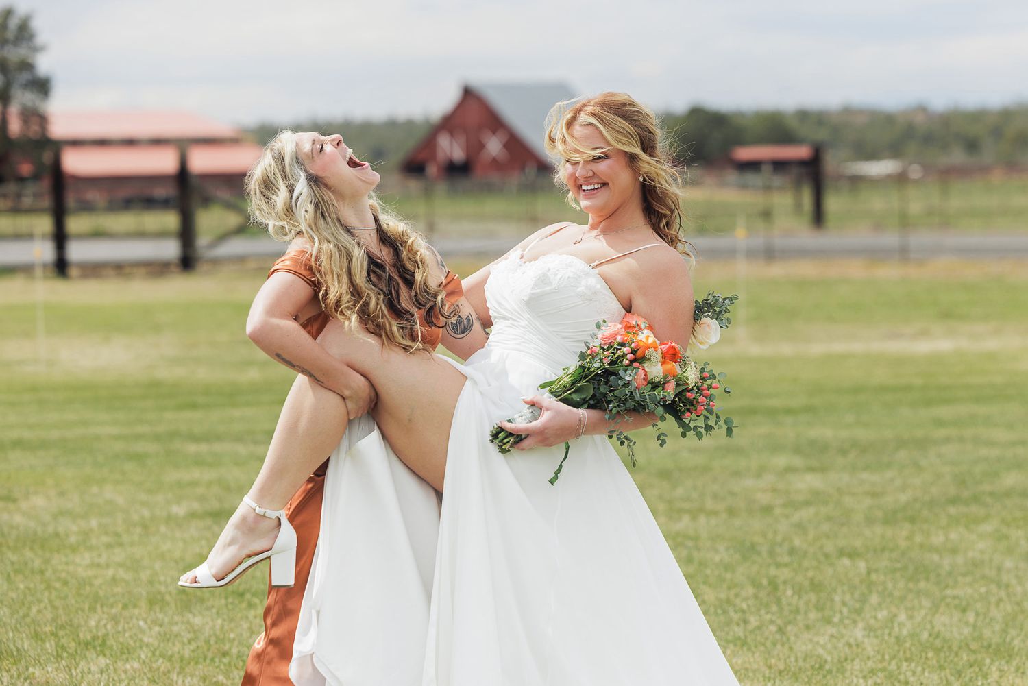 Bride in white gown holds bridesmaid in orange dress outdoors at a farm wedding venue with bouquet.