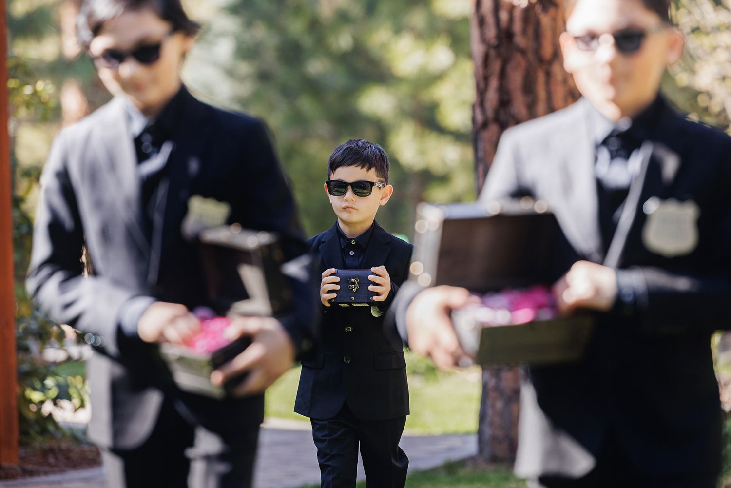 Young boy in black suit and sunglasses holds a gift box at an outdoor wedding ceremony with other boys.