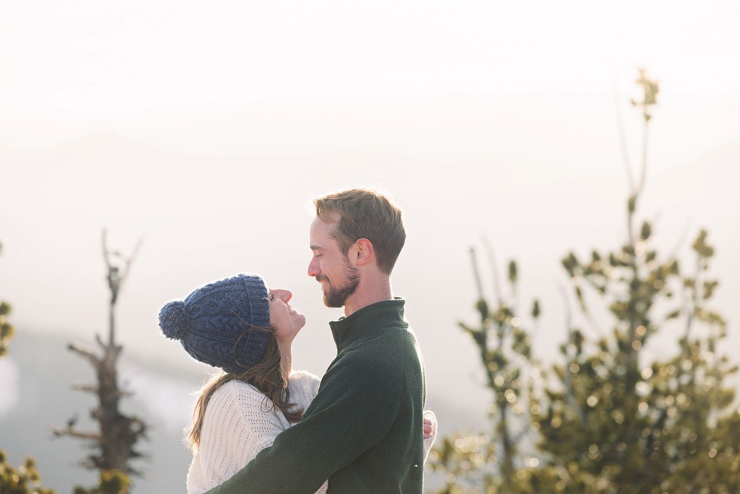 A romantic couple embraces outdoors in winter, the woman wearing a blue knit hat, surrounded by misty mountain scenery.