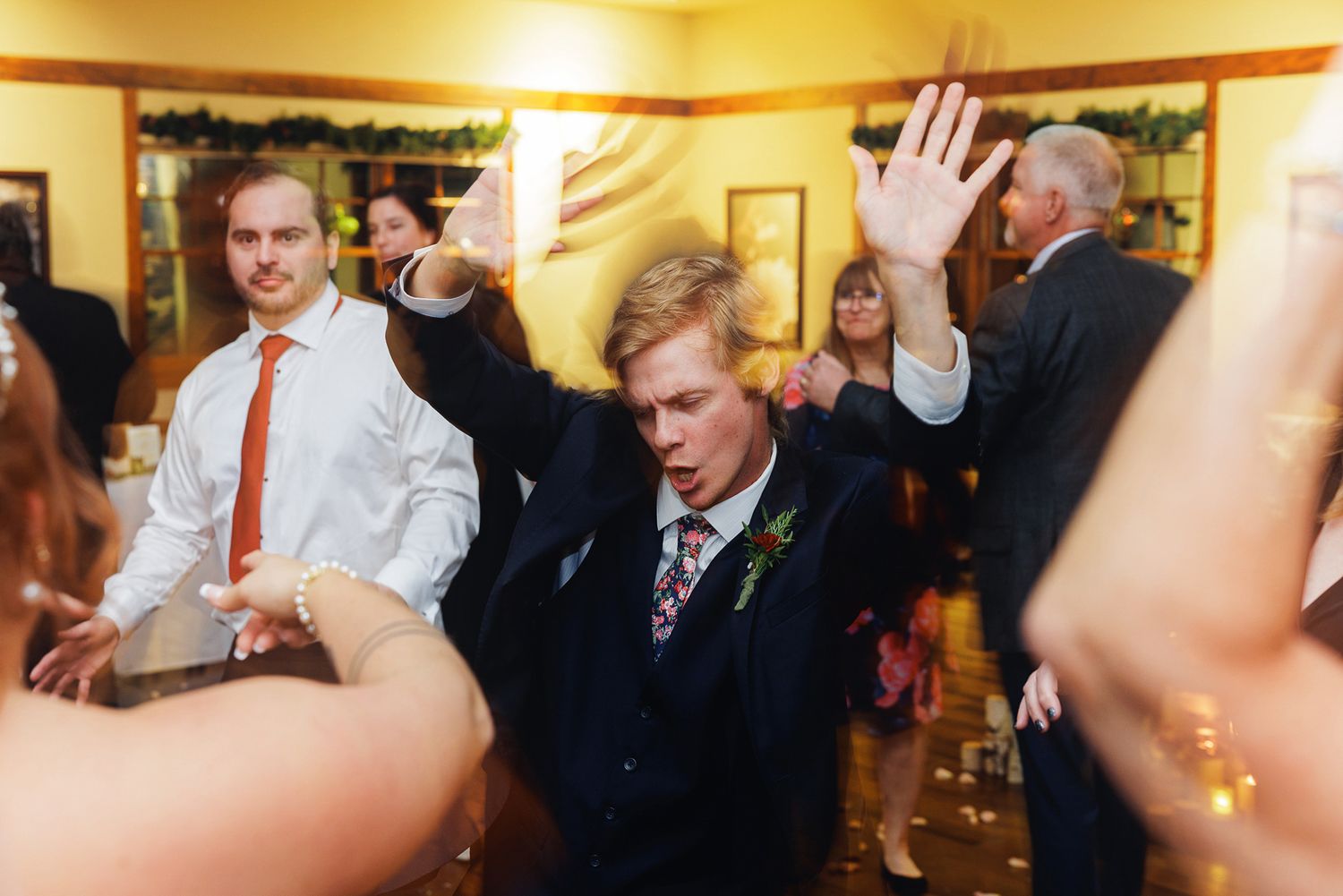 Wedding guests dance with raised hands during a lively reception celebration in a warmly lit indoor venue.