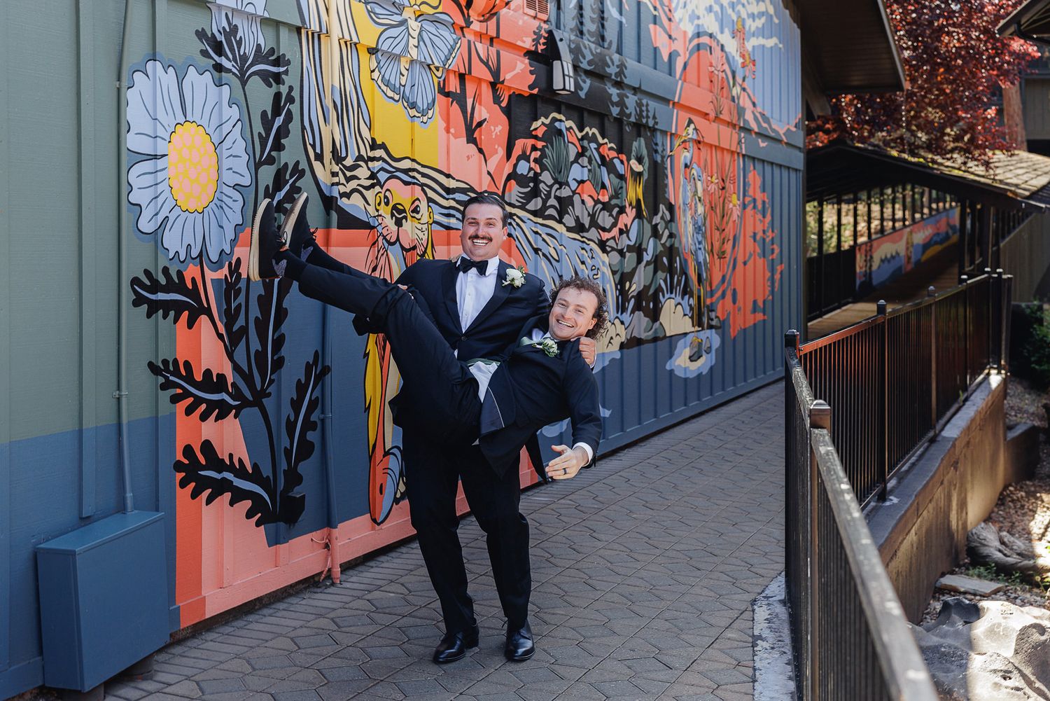 Two grooms in tuxedos pose joyfully in front of a colorful floral mural on their wedding day.