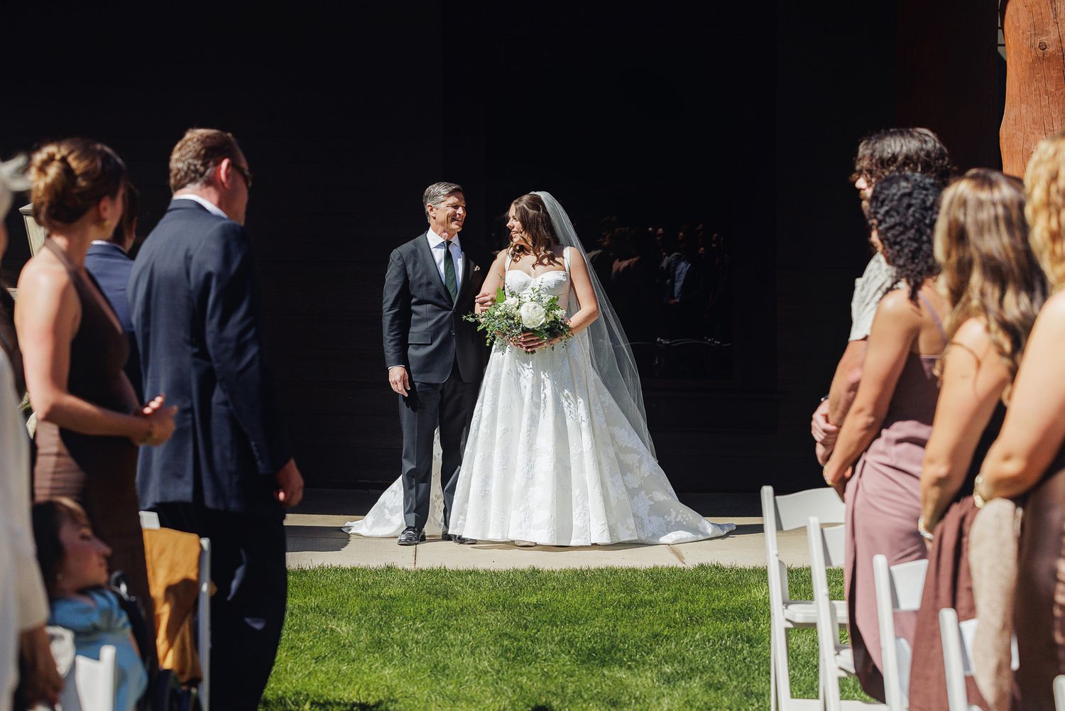 Bride in white ball gown walks down outdoor aisle with escort, guests watching at sunny wedding ceremony.