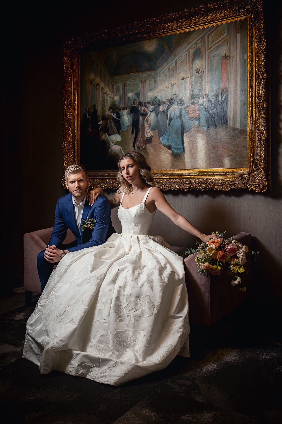 An elegant wedding portrait in front of a classical painting in an ornate gold frame.