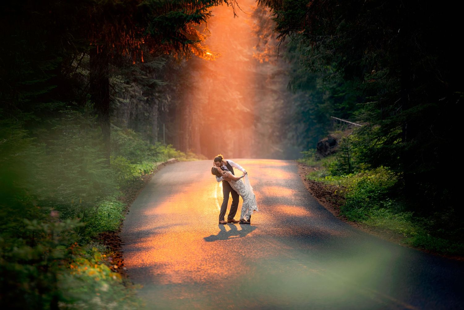 A couple shares an intimate moment on a misty forest road at sunset.