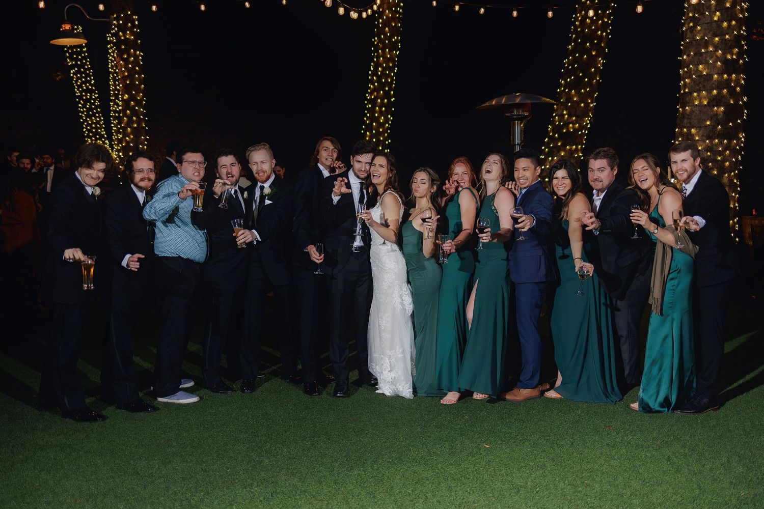 A wedding party poses outdoors at night under twinkling lights with bridesmaids in green dresses and groomsmen in dark suits.