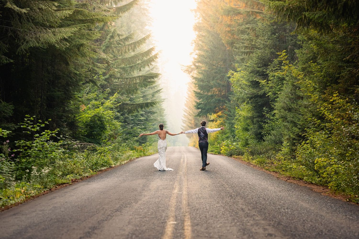 Two figures walk down a tree-lined mountain road surrounded by lush evergreen forest.