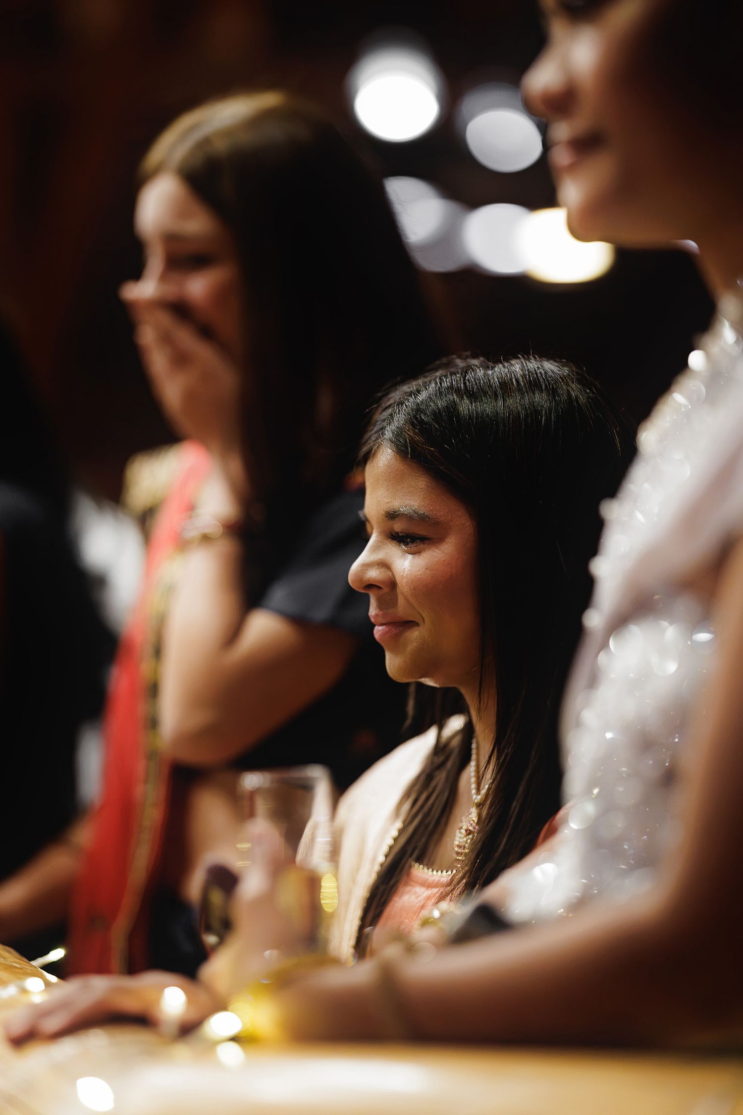 Young girl smiling at a formal event, seated at a table with drinks, surrounded by women in elegant attire.