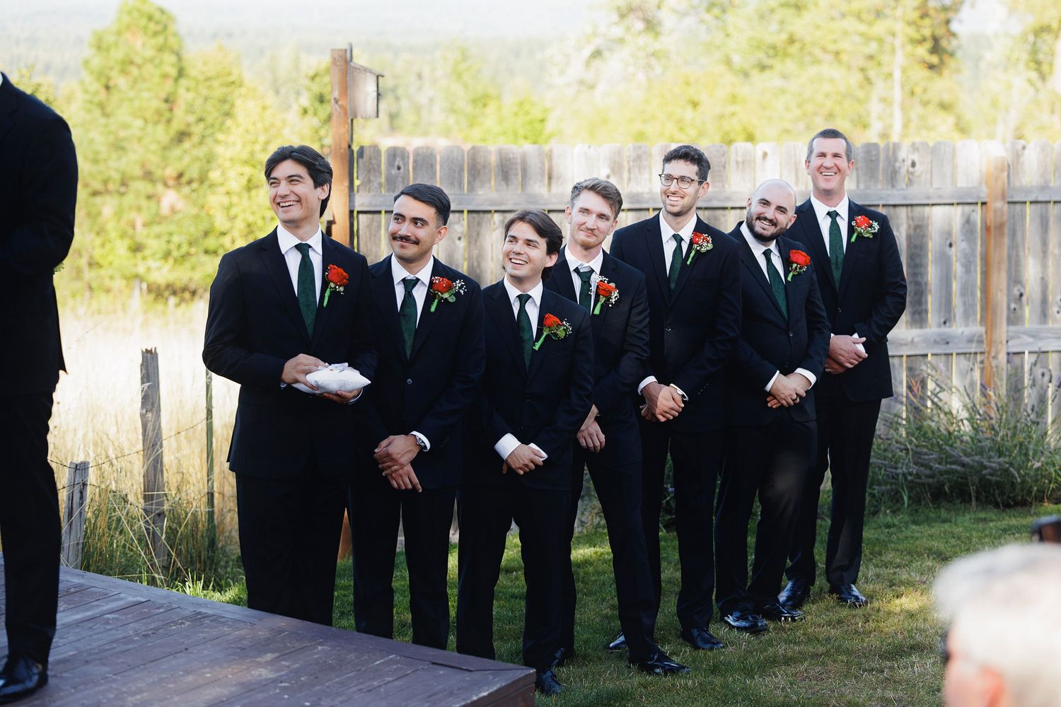 Groomsmen in black suits with red boutonnieres smiling outdoors at a wedding ceremony with rustic wooden fence backdrop.